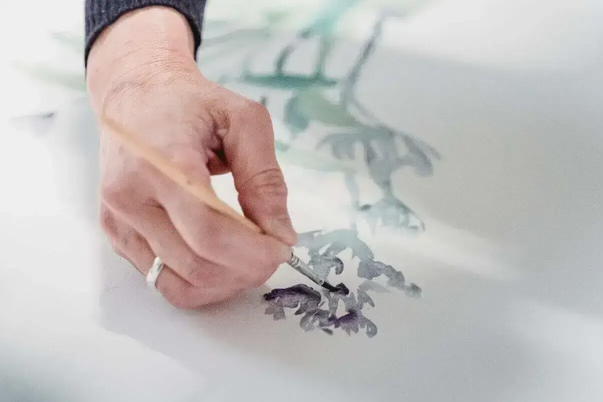 A hand using watercolour to paint a purple flower.